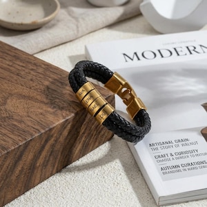May include: A black braided leather bracelet featuring four gold-toned metal cylindrical beads, each engraved with the names AVA, LENA, NAEL, and LOU. The bracelet includes a gold-toned metal clasp mechanism and a double-strand braided leather band.