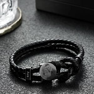 May include: Black braided leather bracelet with a circular metal charm featuring a black and white photo of a couple. The bracelet has a clasp with the name "Ava" engraved on it. The bracelet is a stylish accessory.