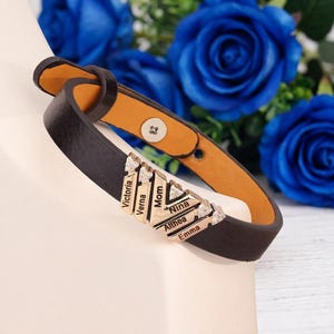 Puede incluir: Una pulsera de cuero marrón oscuro con una pieza central de metal dorado que presenta seis barras verticales grabadas con los nombres Victoria, Verna, Mom, Nina, Althea y Emma, adornada con pequeños cristales claros y un cierre de broche.