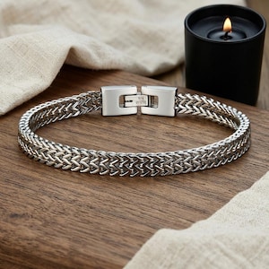 Puede incluir: Una pulsera de acero inoxidable en tono plateado con un diseño de cadena de trigo trenzada. La pulsera incluye un cierre de caja rectangular con un mecanismo de bisagra visible, proporcionando un aspecto seguro y pulido a este accesorio metálico.