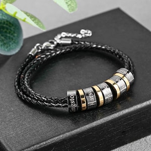 May include: Black braided leather bracelet with silver and gold-toned beads. The beads are engraved with names. The bracelet has a silver clasp. A stylish accessory for everyday wear.