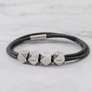May include: Black braided leather bracelet with four silver-colored, geometric beads. Each bead has a name engraved on it. The bracelet has a silver clasp and is displayed on a white marble surface.