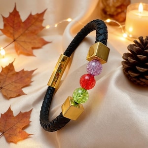 Puede incluir: Una pulsera de cuero trenzado negro con un cierre cilíndrico dorado grabado con el texto I Love You, dos cuentas cúbicas doradas y tres cuentas redondas de vidrio craquelado en colores verde claro, rojo y morado claro.