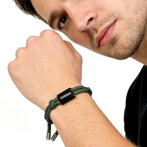 May include: Olive green bracelet with a black rectangular clasp that reads "custom" in white lettering. The bracelet is made of braided cord and features silver-colored metal end pieces. The bracelet is worn on a wrist.