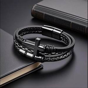 May include: Black leather bracelet featuring a cross pendant. The bracelet has multiple strands with a braided design and metal accents. The cross is centered on the bracelet, and the clasp is magnetic.