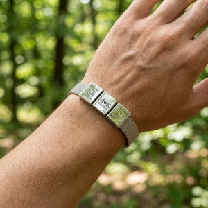 May include: A silver-toned bracelet featuring three square metal charms. The center charm is engraved with the letters M and K separated by a heart and the date 13.03.2016. The two outer charms feature engraved baby illustrations and text.