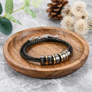 May include: A black braided leather bracelet featuring multiple cylindrical metal beads. Each bead is engraved with a name, including Kingston and Karson, and is accented with gold-toned rings. The bracelet includes a silver-toned metal chain clasp.