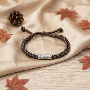 May include: A dark brown braided cord bracelet featuring five cylindrical silver-toned beads. The beads are engraved with small symbols, a heart, and the text "ame" and "Sara". The bracelet has an adjustable sliding knot closure.