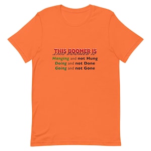 May include: Orange t-shirt with the text "THIS BOOMER IS Hanging and not Hung Doing and not Done Going and not Gone" in red and green. The shirt is a short-sleeved crew neck.