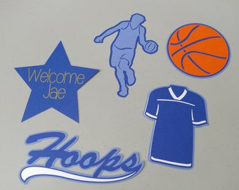 May include: Blue and white basketball themed party decorations, including a star with the text "Welcome Jae", a basketball, a jersey, and the word "Hoops" in a stylized font.