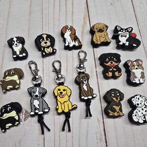 May include: A collection of dog-themed charms and keychains. The charms feature various dog breeds in different colors and poses. Some are attached to silver-colored keychains with black cords. The background is a light wood-grain pattern.