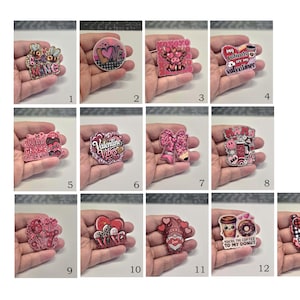 May include: A collection of thirteen acrylic badge reels featuring Valentine's Day themes, including hearts, gnomes, cows, bows, and coffee motifs with text like 'Be Mine', 'Valentine Vibes', 'Mama', and 'You're the coffee to my donut' in pink, red, and black.