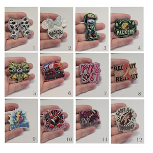May include: A collection of twelve decorative acrylic badges featuring various sports and awareness themes, including Green Bay Packers logos, football graphics, pink awareness ribbons, and text such as Pink Out, Red Out, and Game Day Touchdown.