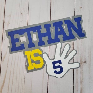 May include: Gray, blue, and yellow paper cutouts that spell out "Ethan is 5" with a white hand cutout with a blue "5" on it.
