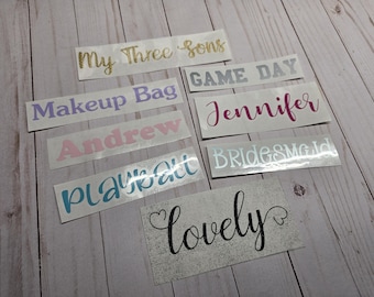 May include: A collection of adhesive vinyl decals featuring various words and names in different fonts and colors, including My Three Sons in gold, GAME DAY in silver, Makeup Bag in purple, Jennifer in magenta, Andrew in pink, playball in blue, BRIDESMAID in light blue, and lovely in black with heart accents on a silver glitter background.