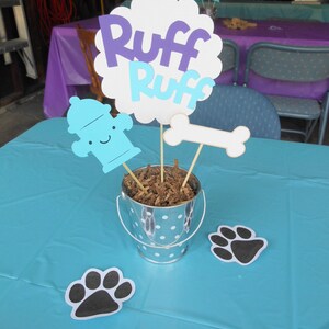 May include: A blue and purple "Ruff Ruff" sign with a fire hydrant, bone, and paw print decorations on a blue tablecloth. The decorations are in a silver bucket with white polka dots and brown shredded paper.