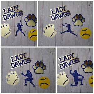 May include: A collage of softball-themed cutouts. The words "LADY DAWGS" are in gold and navy blue. Other cutouts include a paw print, a softball, a glove, and a softball player in navy blue. The items are arranged on a light wood-grain surface.