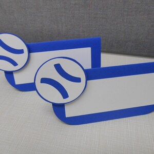 May include: Two blue and white place card holders with a baseball design. Each card holder has a white rectangle for writing a name and a white baseball with blue stitching.