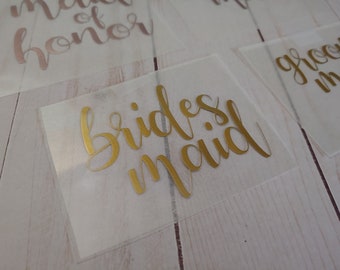 May include: Several rectangular, translucent adhesive decals featuring metallic gold and rose gold calligraphy text. The visible labels display the words bridesmaid, maid of honor, and groomsman in a flowing, cursive script font style.