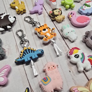 May include: A collection of colorful animal-shaped charms, including a tiger, dinosaur, and koala bear, with silver-tone keychains. Other charms include a hedgehog, deer, and a pink flamingo. The charms are made of rubber and are on a light wood surface.