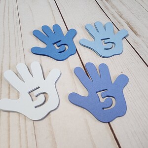 May include: Four paper hand cutouts, two white and two blue, with the number "5" cut out of the center of each hand. The hands are arranged in a square pattern on a light wood surface.