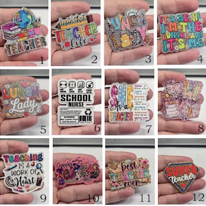 May include: A collection of twelve glittery acrylic pins featuring school-themed designs and text, including phrases like Teacher, Livin that Teacher life, Lunch Lady, School Nurse, She is a Teacher, Teaching is a work of heart, and Super Teacher.