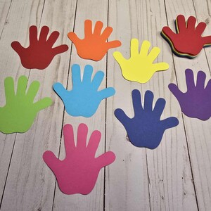 May include: A collection of colorful hand-shaped cutouts in various shades, including red, orange, yellow, green, blue, purple, and pink. The cutouts are arranged on a light-colored wooden surface.