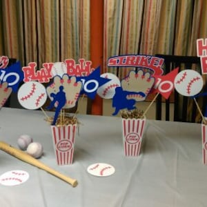 May include: Five baseball-themed centerpieces with red, white, and blue decorations. Each centerpiece features a popcorn container with red and white stripes, a baseball, and various cutouts with baseball-related words and images, including "All Star", "Play Ball", "Strike", and "Home Run".