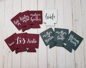 May include: A collection of personalized drink koozies in various colors, including burgundy, white, and teal. Each koozie features white script text with phrases like "Bride," "Bridesmaid," and names, perfect for a wedding or bridal party.