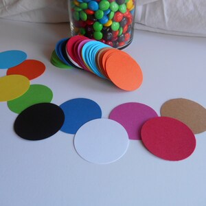 May include: A collection of colorful, oval-shaped paper cutouts in various colors, including blue, orange, pink, and brown. A glass jar filled with colorful candies is in the background. The cutouts are arranged on a white surface.