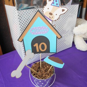May include: A blue and brown dog house with the name Allison and the number 10 on it. The dog house is sitting on a white polka dot bucket with brown shredded paper inside. There is a white bone and a blue dog bowl with brown food inside.