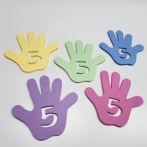 May include: Five colorful hand-shaped cutouts in yellow, green, blue, purple, and pink. Each hand has the number 5 cut out in the center. The cutouts are arranged on a white surface.