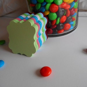 May include: A stack of colorful paper tags with scalloped edges. The tags are in shades of green, blue, pink, and white. There are also some colorful candies in the background.