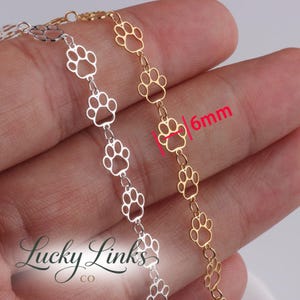 Puede incluir: Primer plano de dos cadenas con recortes de huellas de pata. Una cadena es plateada, la otra dorada. Los recortes de huellas de pata miden 6 mm de ancho. El logotipo de Lucky Links Co. es visible en la esquina inferior izquierda.