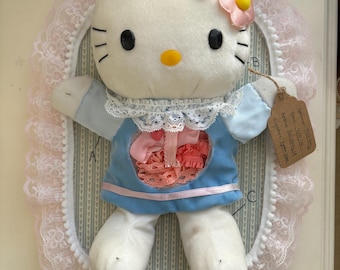 Op de afbeelding: Een wit Hello Kitty pluche speelgoed met zwarte ovale ogen en een gele neus. Het draagt een blauwe jurk met een witte kanten kraag en roze bloemhaaraccessoires. Een cirkelvormige uitsnede op de jurk onthult roze stof. Een bruin papieren label zit vast.