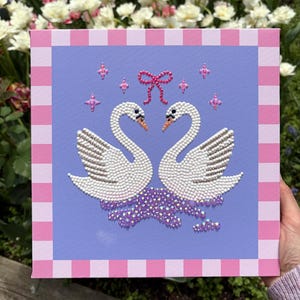 May include: Square canvas art featuring two white swans facing each other, embellished with small white beads and silver accents. A pink bow and star-shaped embellishments are above the swans. The canvas has a light purple background and a pink and white checkered border.