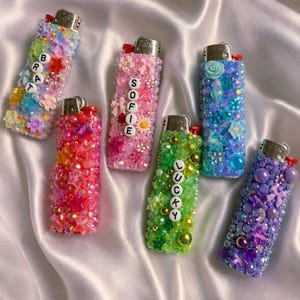 May include: Six rectangular lighters decorated with colorful beads, small plastic flowers, stars, and bows. Four lighters feature white letter beads spelling out BRAT, SOFIE, and LUCKY. The lighters are arranged in a cluster, each with a metallic top.