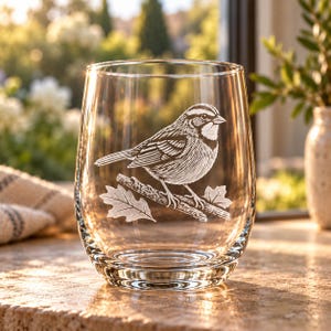 May include: A clear, stemless glass tumbler featuring a white etched illustration of a small bird perched on a branch with two oak leaves. The design is centered on the front of the glass, showcasing detailed line work of the bird's feathers.