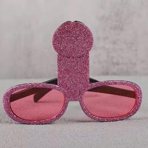 May include: Novelty sunglasses with a pink glitter penis-shaped frame and pink lenses. The sunglasses have a black bridge and arms. The frame is a solid pink color with glitter.