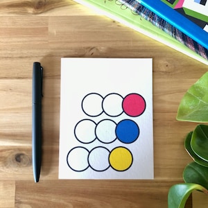 May include: A white card with a graphic design of circles in black outlines, with three circles filled in with pink, blue, and yellow. A black pen is to the left of the card. Books are in the background.