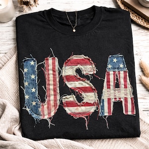 May include: Black t-shirt with the word "USA" appliqued on the front. The letters are made from denim and red and white striped fabric, resembling the American flag. The letters have a distressed, vintage look.
