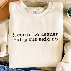 Op de afbeelding: Cr&egrave;mekleurige sweatshirt met de zwarte tekst "i could be meaner but jesus said no". De sweatshirt heeft een ronde hals en lange mouwen. Het kledingstuk is opgevouwen en lijkt van een zacht materiaal te zijn.