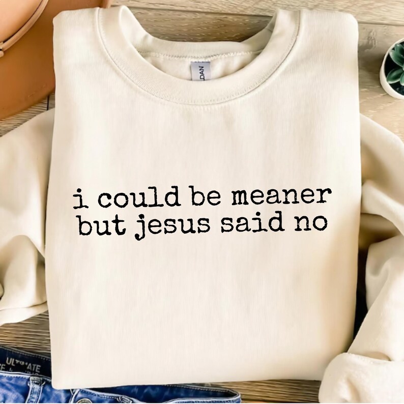 Op de afbeelding: Cr&egrave;mekleurige sweatshirt met de zwarte tekst "i could be meaner but jesus said no". De sweatshirt heeft een ronde hals en lange mouwen. Het kledingstuk is opgevouwen en lijkt van een zacht materiaal te zijn.