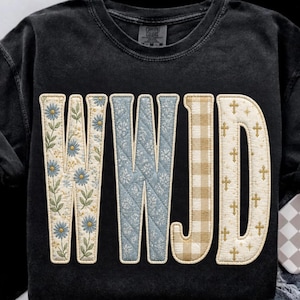May include: A black crewneck sweatshirt featuring the letters WWJD in a large, quilted-style font. The Ws have a blue floral pattern, the J has a tan gingham check, and the D is cream with a gold cross and star pattern.