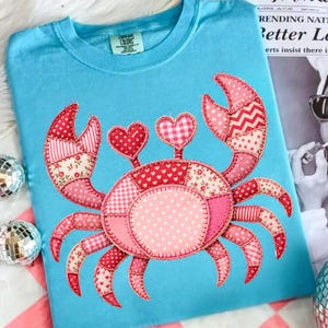 May include: A light blue t-shirt featuring a patchwork crab design in shades of pink and red. The crab has heart-shaped eyes and is made of various patterned fabrics. The shirt has a crew neck and short sleeves.