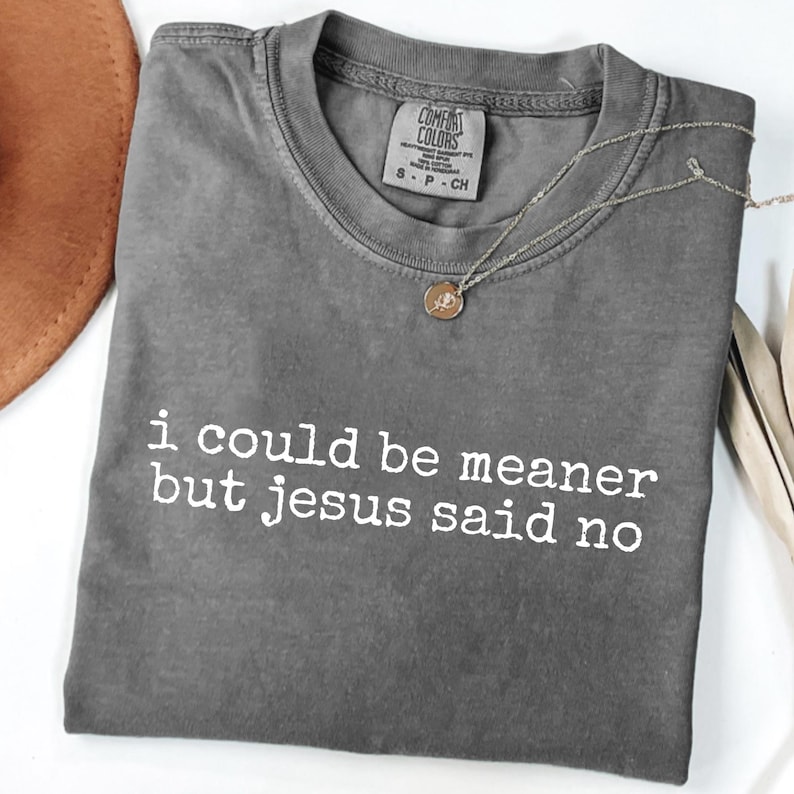 Op de afbeelding: Een grijs Comfort Colors T-shirt met de witte tekst "i could be meaner but jesus said no". Een gouden hangerketting en een bruine hoed zijn ook zichtbaar.