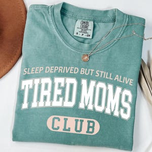 May include: A teal Comfort Colors t-shirt with the text "SLEEP DEPRIVED BUT STILL ALIVE TIRED MOMS CLUB" in white and pink lettering. A silver necklace with a pendant is draped across the shirt.