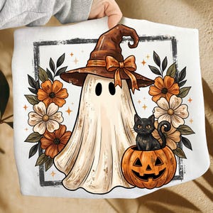 May include: A white sweatshirt featuring a graphic print of a ghost wearing a brown witch hat with an orange bow, sitting next to a black cat inside an orange jack-o'-lantern, surrounded by orange and white flowers and small star accents.