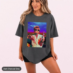 May include: A dark charcoal grey short-sleeve t-shirt featuring a vibrant, multi-coloured graphic print of a person with blonde hair wearing sunglasses, holding a cigarette and a bottle against a sunset-toned sky and cityscape background.