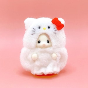 May include: A small, fuzzy white figurine wearing a hooded costume that resembles a cat character. The hood features black embroidered eyes, a yellow nose, and a bright red bow on the side. The figurine has a round, soft texture throughout.